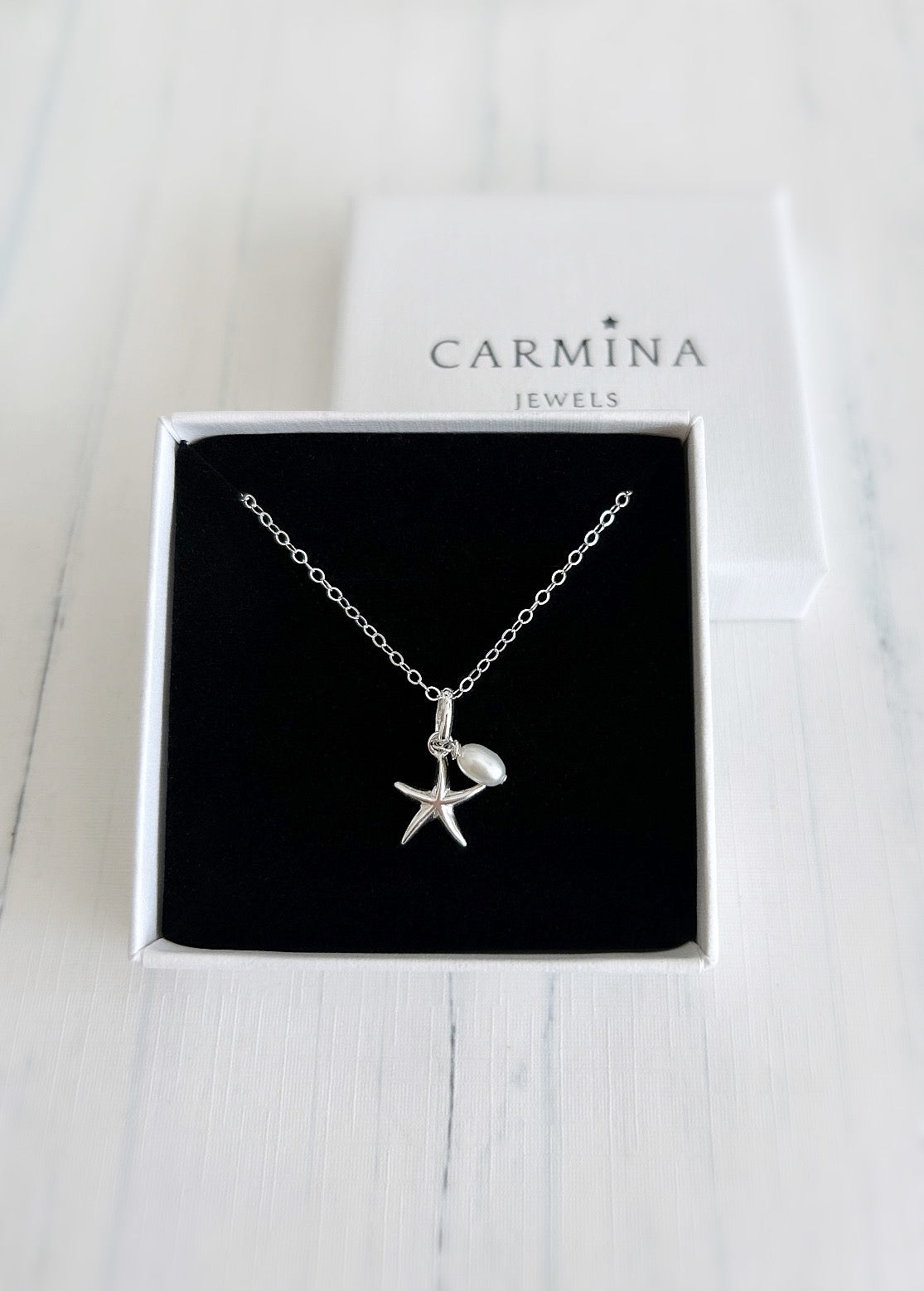 A silver starfish pendant with a pearl charm, presented in a box with the Carmina Jewels branding.