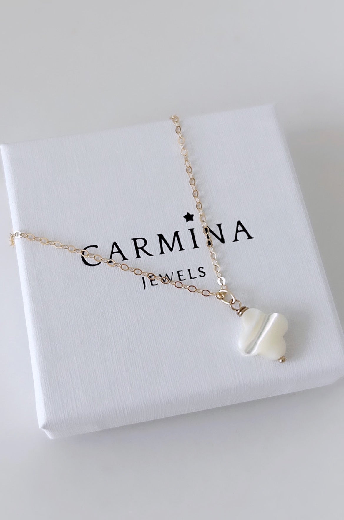 A Mother of Pearl clover shaped pendant necklace in a white and gold color, displayed on a delicate 16” cable chain, presented in a 'Carmina' branded gift box.