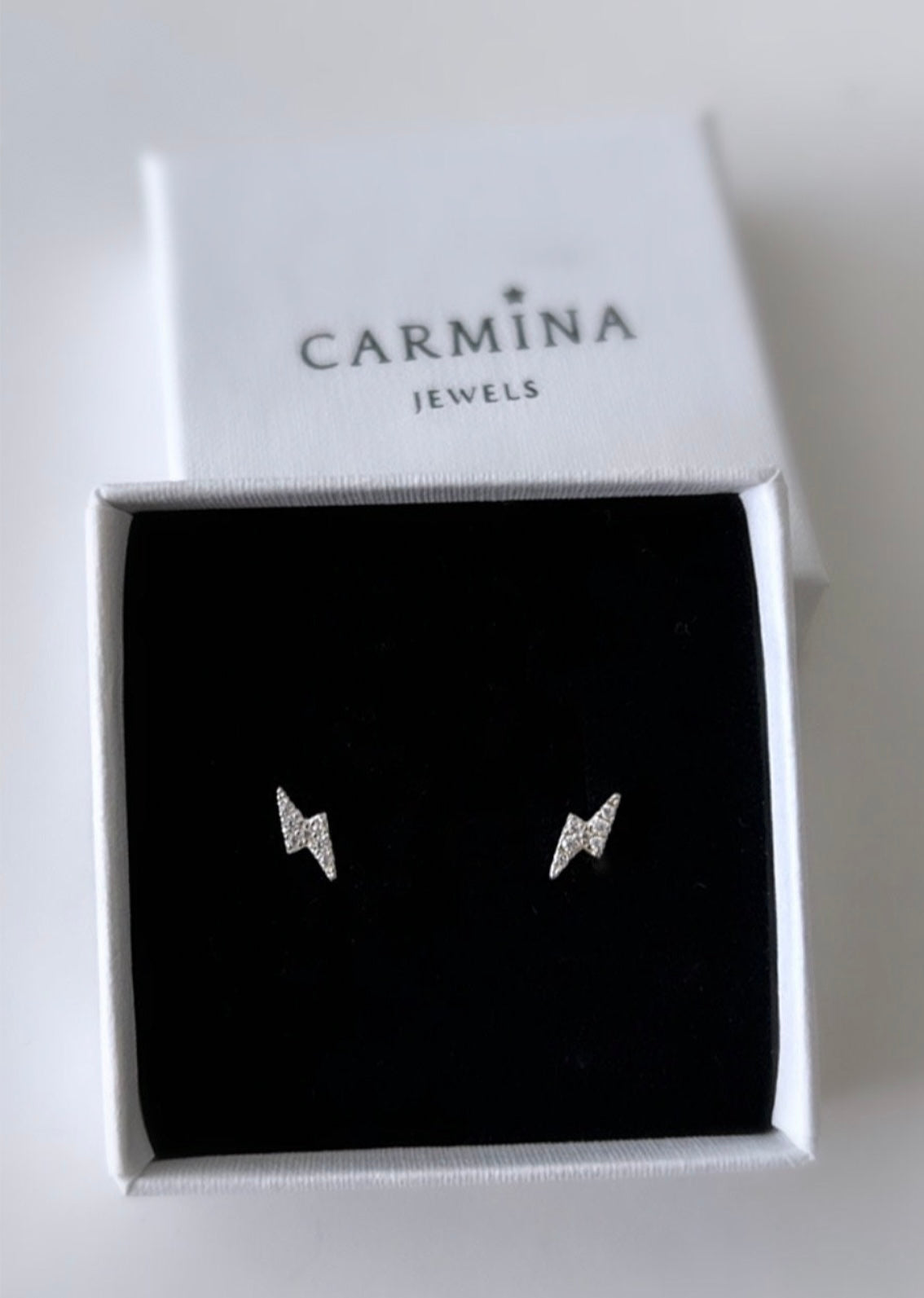 A pair of silver lightning bolt stud earrings encrusted with cubic zirconia crystals,  in a white gift box with 'Carmina Jewels' branding.