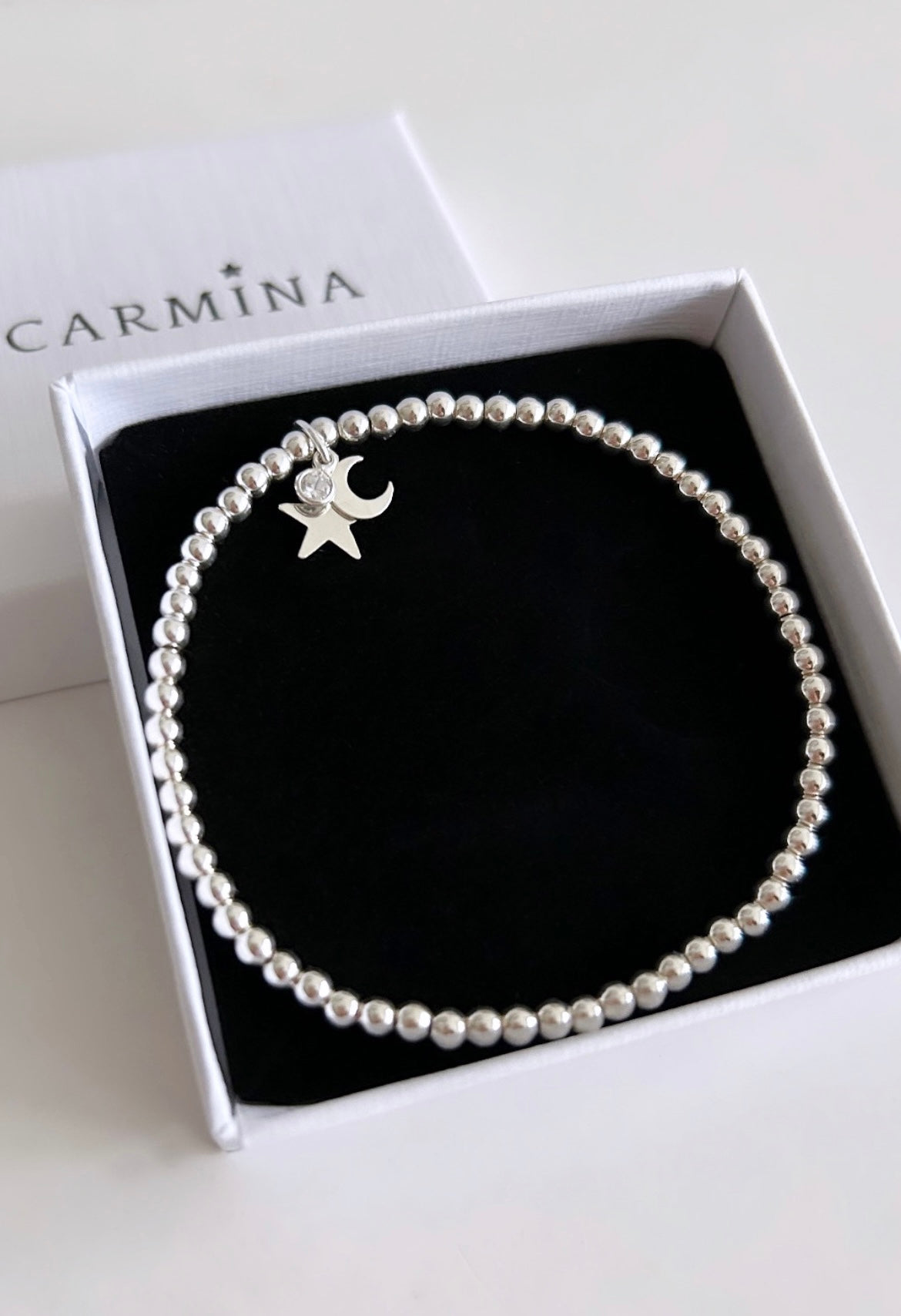 A sterling silver charm bracelet with a crescent moon and star charm, presented in a  gift box with 'Carmina' branding.