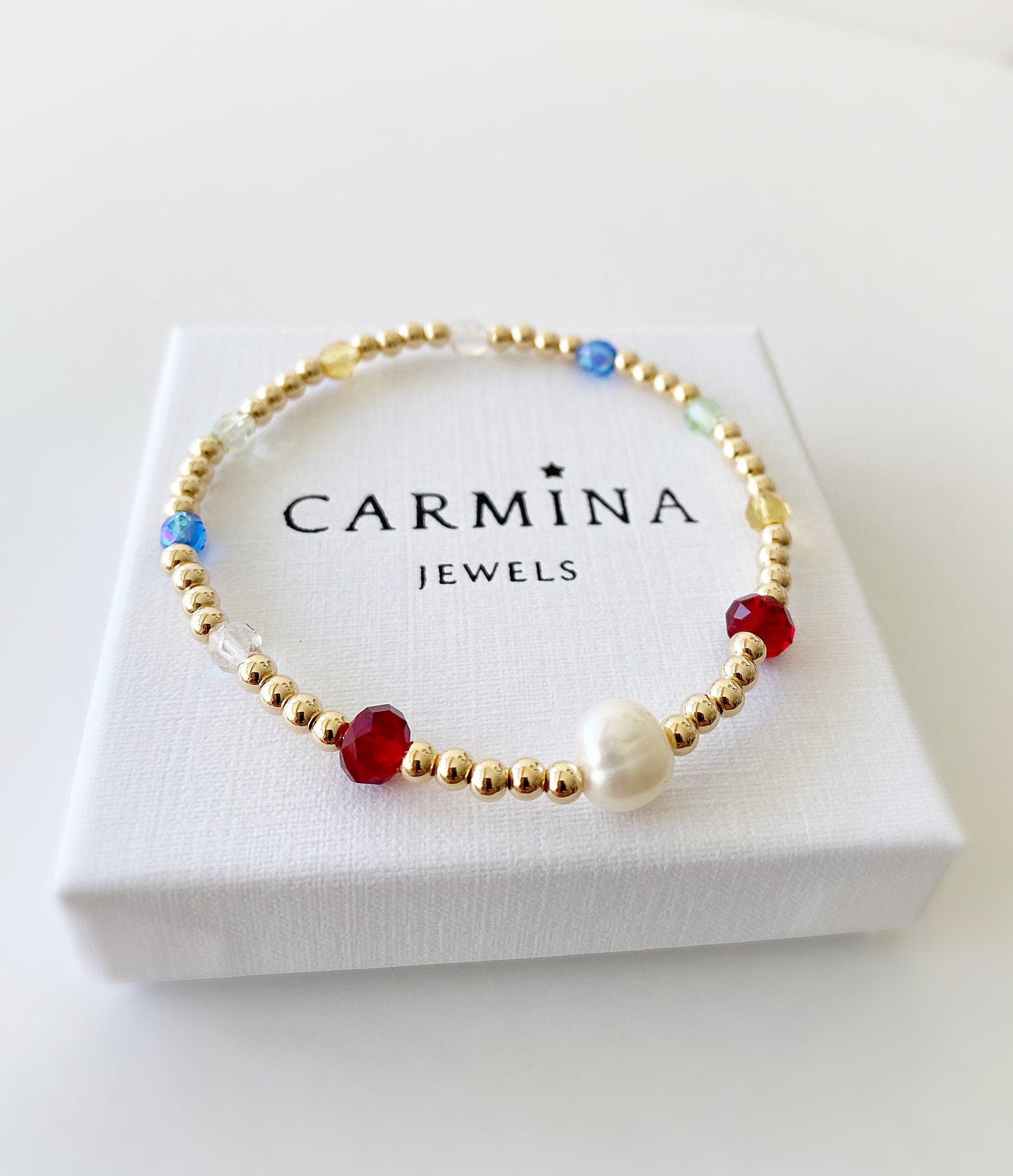 A gold-colored bracelet with blue, red, and white glass beads, featuring a central baroque freshwater pearl, presented in a white box with 'CARMINA JEWELS' branding.