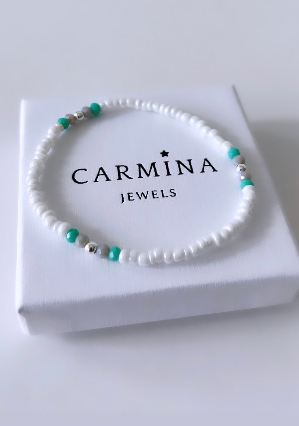 A seed bead bracelet with aqua and pale grey crystal glass beads, and gold/silver spacer beads presented in a white box with 'CARMINA JEWELS' branding.