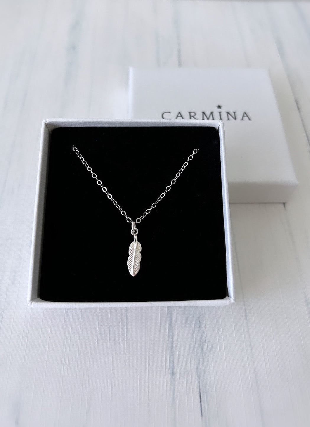 A silver feather pendant necklace in a gift box with 'Carmina' branding.