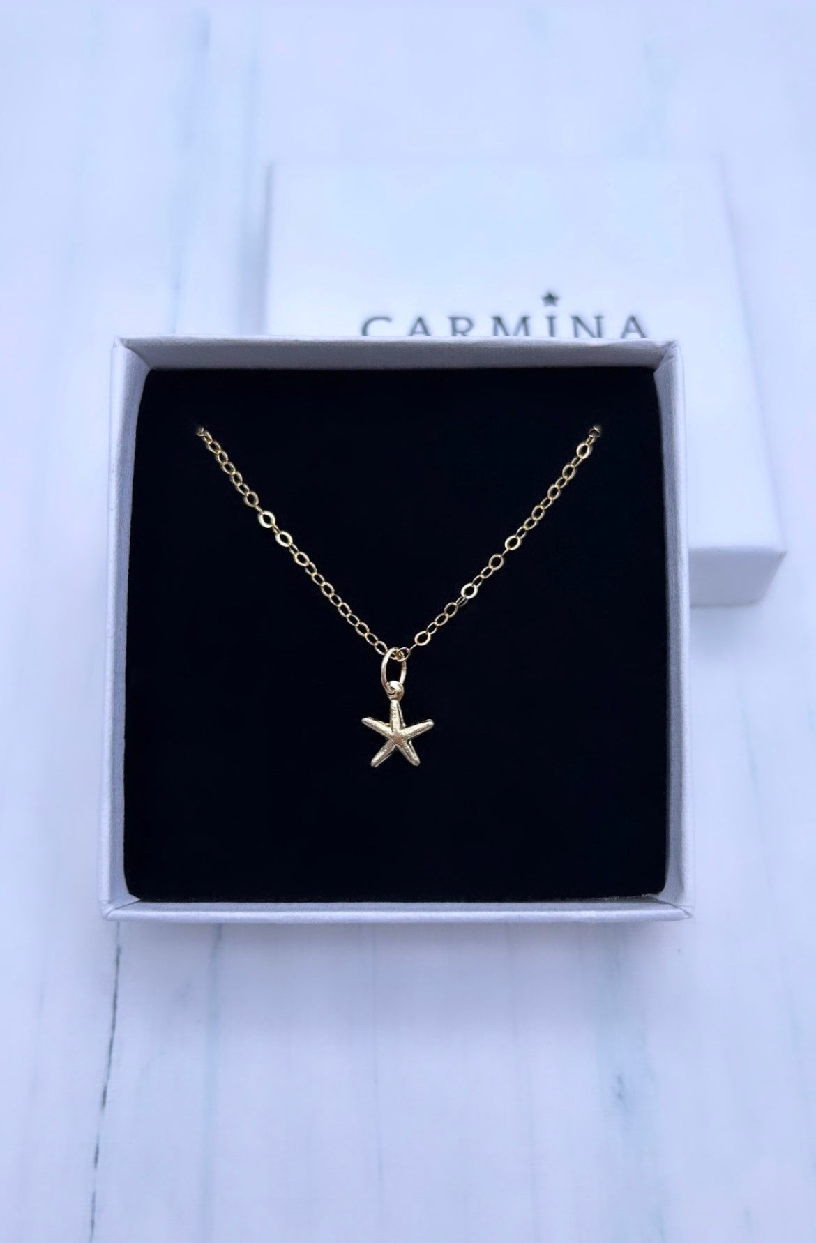 A gold-filled starfish pendant necklace presented in a black and white box with 'CARMINA' branding.